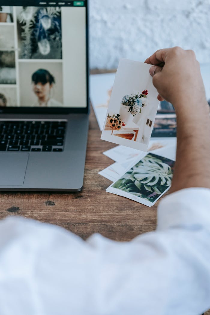 Hand holding photo near laptop in creative workspace, showcasing artistic inspiration.
